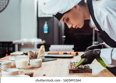 side view of chocolatier in latex gloves holding pastry bag with caramelized nuts near chocolate molds