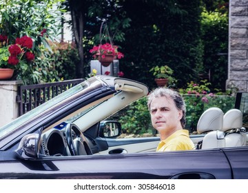 Side View Of Classy  40 Years Old Sportsman With Three-day Beard And Salt And Pepper Hair Wearing A Yellow Polo Shirt While He Is Driving A Dark Brown Car In Residential Neighborhood