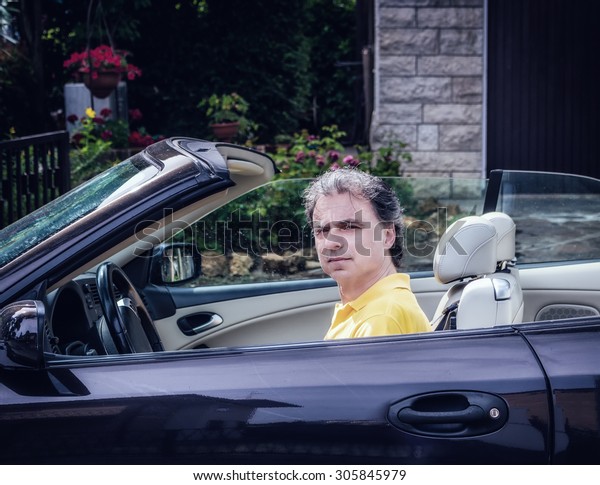 Side
view of classy  40 years old sportsman with three-day beard and
salt and pepper hair wearing a yellow polo shirt while he is
driving a dark brown car in residential
neighborhood