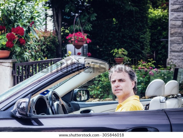 Side
view of classy  40 years old sportsman with three-day beard and
salt and pepper hair wearing a yellow polo shirt while he is
driving a dark brown car in residential
neighborhood