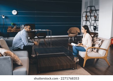Side view of client having difficult situation sitting on sofa talking to psychotherapist during therapy consultation getting help from sessions