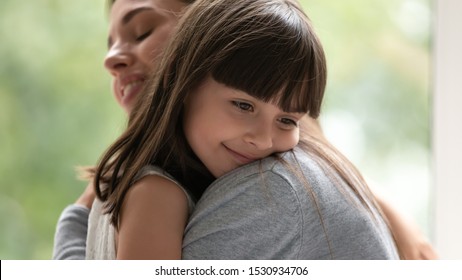 Side view close up head shot happy little adopted kid girl put head on mothers shoulder, feeling love and support. Small cute daughter hugging embracing cuddling young smiling mother at new home.