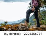 Side view, Close up legs of young asian hiking man standing, camera on kneck and holding water drink bottle with happy on peak of rocky mountain, beautiful landscape in background, copy space