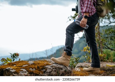 Side view, Close up legs of young asian hiking man standing, camera on kneck and holding water drink bottle with happy on peak of rocky mountain, beautiful landscape in background, copy space