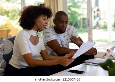 Side View Close Up Of A Mixed Race Couple Sitting In The Living Room On The Couch, Looking At Paperwork And Using A Laptop Computer Together