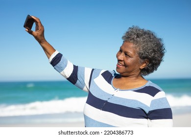 Side view close up of a senior African American woman standing on the beach with blue sky in the background, smiling and taking a selfie with her smartphone - Powered by Shutterstock