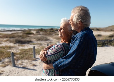 Side view close up of a senior Caucasian couple at the beach in the sun, standing and embracing, leaning against their car, smiling and admiring the view