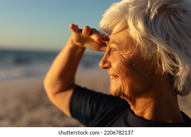 Side view close up of a senior Caucasian woman at the beach at sunset, shielding her eyes from the sun, smiling and looking out to sea - Powered by Shutterstock