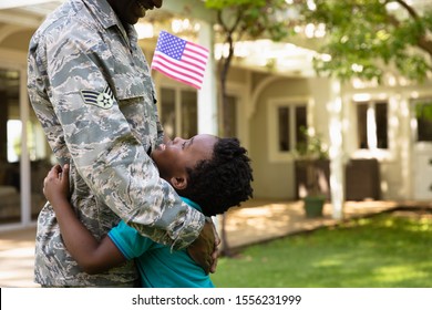 Side View Close Up Of A Young Adult African American Male Soldier In The Garden Outside His Home, Embracing His Young Son, Who Is Looking Up At Him Smiling And Holding A US Flag, Thier House In The