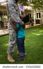 SIde View Close Up Of A Young Adult African American Male Soldier In The Garden Outside His Home, Embracing His Young Son, Who Is Looking Up At Him Smiling And Holding A US Flag, Thier House In The