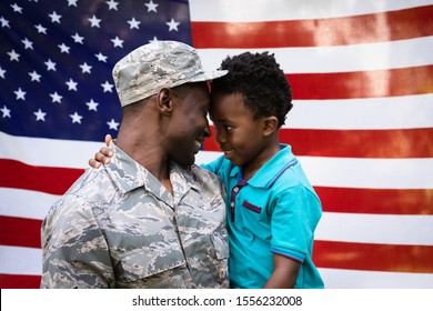 Side View Close Up Of A Young Adult African American Male Soldier Carrying His Young Son, Both Looking At Each Other And Smiling, In Front Of A US Flag