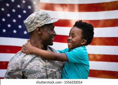 Side View Close Up Of A Young Adult African American Male Soldier Carrying His Young Son, Both Looking At Each Other And Smiling, In Front Of A US Flag