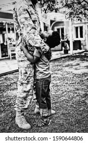 SIde View Close Up Of A Young Adult African American Male Soldier In The Garden Outside His Home, Embracing His Young Son, Who Is Looking Up At Him Smiling And Holding A US Flag, Thier House In The