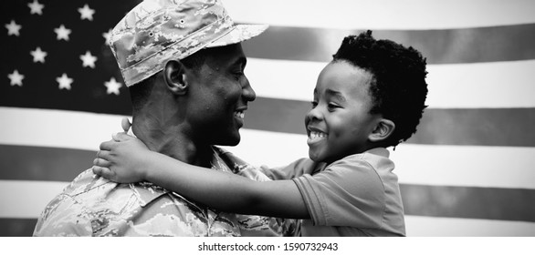 Side View Close Up Of A Young Adult African American Male Soldier Carrying His Young Son, Both Looking At Each Other And Smiling, In Front Of A US Flag