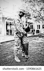 SIde View Close Up Of A Young Adult African American Male Soldier In The Garden Outside His Home, Embracing His Young Son, Who Is Looking Up At Him Smiling And Holding A US Flag, Thier House In The