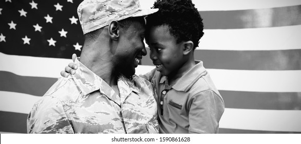 Side View Close Up Of A Young Adult African American Male Soldier Carrying His Young Son, Both Looking At Each Other And Smiling, In Front Of A US Flag