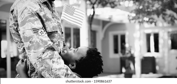 Side View Close Up Of A Young Adult African American Male Soldier In The Garden Outside His Home, Embracing His Young Son, Who Is Looking Up At Him Smiling And Holding A US Flag, Thier House In The