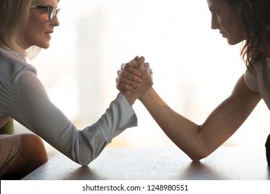 Side view close up young brunette and aged blond businesswomen arm wrestling holds elbows on table, exerting pressure on each other, looking eyes to eyes face to face struggling for leadership at work