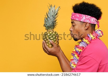 Side view close up young fun cool cheerful man 20s he wear pink t-shirt hawaiian lei near hotel pool hold pineapple fruit isolated on plain yellow background studio. Summer vacation sea rest concept
