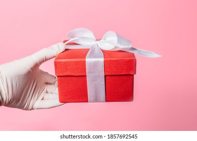 Side View Closeup Of Human Hand In White Surgical Gloves Holding A Red Gift Box Isolated On Pink Background. Studio Shot