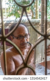 Side View Close-up Portrait Of Senior Indian Man Shaving Beard With A Razor In Front Of A Small Mirror. 