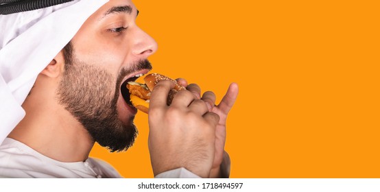 Side View Of Close-up Portrait Of Young Arab Man Eating A Burger. Hungry Arabic Guy Isolated On Orange Background Eating A Hamburger. Man Wearing Kandura And Ghutra Eating Junk Food.