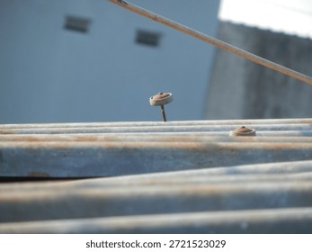 Side view close-up of a rusty nail protruding from an old corrugated metal roof sheet. Selective focus on the weathered fastener against a blurred building wall background