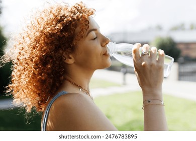 Side view closeup of thirsty athletic redhead female drinking pure water from transparent bottle after workout training outdoors, quenches her thirst with closed eyes, having rest after yoga practice - Powered by Shutterstock