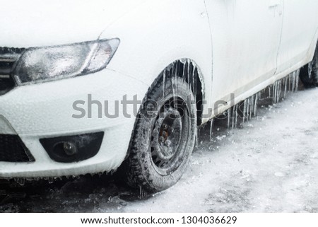 side view closeup of white frozen car with freezing rain icicles hanging on tires stopped in a parking lot covered in snow winter time