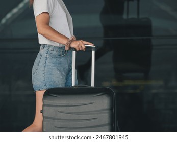 Side view close-up of a woman in casual attire holding the handle of a black suitcase outside an airport terminal with reflective glass windows. Travel and holidays preparation scene - Powered by Shutterstock