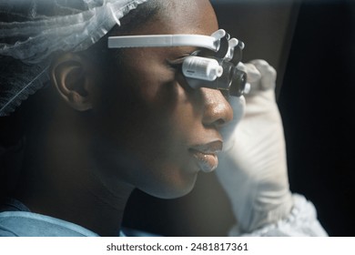 Side view closeup of young African American woman as female surgeon wearing surgical loupe in hazy operating room preparing for microsurgery copy space - Powered by Shutterstock