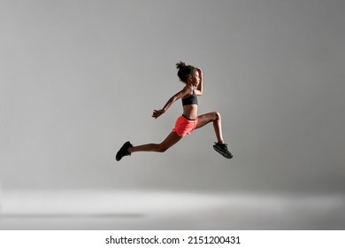 Side view of concentrated athletic black girl jumping during running. Female child wear sportswear. Modern healthy and sports youngster lifestyle. Isolated on grey background. Studio shoot. Copy space