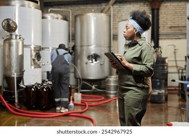 Side view of concentrated female African American process technician wearing denim overall inspecting tanks with craft cider while using tablet computer in workshop of factory, copy space - Powered by Shutterstock