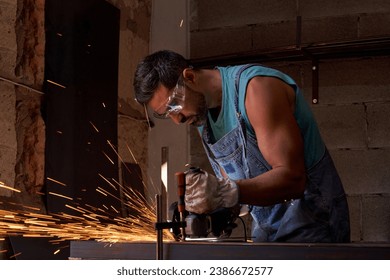 Side view of concentrated man wearing workwear and protective goggles standing at workbench and cutting sparking metal while using professional equipment in metalwork workshop - Powered by Shutterstock