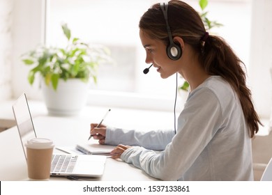 Side view concentrated millennial mixed race woman wearing headset with microphone, looking at laptop screen, listening educational webinar, taking notes, getting remote online education at home.