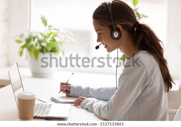 Side view concentrated millennial mixed race woman\
wearing headset with microphone, looking at laptop screen,\
listening educational webinar, taking notes, getting remote online\
education at home.
