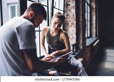 Side view of concentrated muscular male in white shirt sharing tablet with pretty young female while sitting and resting on windowsill - Powered by Shutterstock