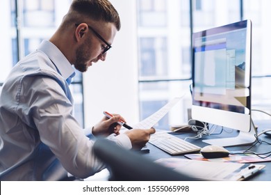 Side view of concentrated smart specialist verifying document and making note with pen working at table with computer in light office - Shutterstock ID 1555065998