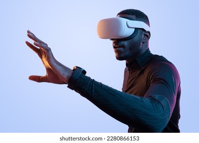 Side view of concentrated young African American man with dark hair and beard in black athletic clothes, and VR glasses touching air while exploring virtual reality in neon studio - Powered by Shutterstock