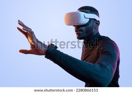 Side view of concentrated young African American man with dark hair and beard in black athletic clothes, and VR glasses touching air while exploring virtual reality in neon studio