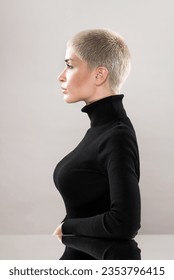 Side view of concentrated young female with short blond hair in black turtleneck sitting at mirrored table and looking away with confidence against beige background - Shutterstock ID 2353796415