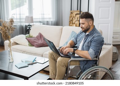 Side View Of Concentrated Young Man In Wheelchair Typing On Wireless Laptop In Bright Living Room. Caucasian Male Employee With Disability Working At International Company From Home.
