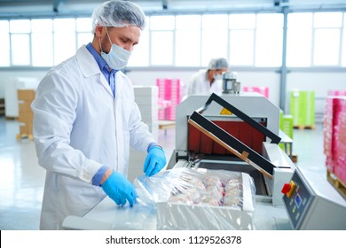 Side view of confectionery factory worker in white coat using machinery to pack paper box with pastry into plastic film.