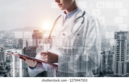Side view of confident female doctor in white sterile coat making notes in notebook while standing outdoors with city view and medical interface icons on background. Medical industry. Double exposure