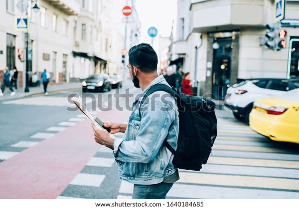 Side view of confident male traveler in denim and\
with backpack checking location with cellphone and map while\
standing in urban street
