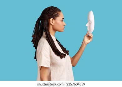 Side view of confident serious woman with dreadlocks holding and looking at white mask with attentive look, trying to understand hiding personality. Indoor studio shot isolated on blue background.