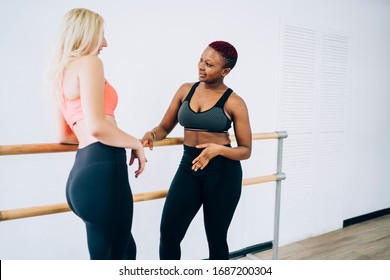 Side View Of Confident Toned Diverse Women In Sportswear Standing Face To Face In Bright Fitness Center Exercise Room And Talking Seriously While Leaning On Ballet Barre