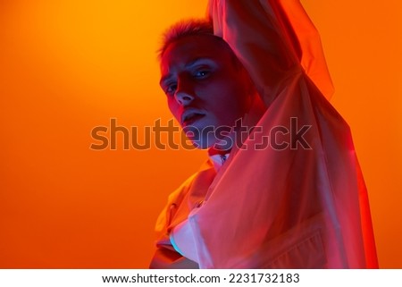 Side view of confident young extravagant female model with short blond hair in translucent coat looking at camera against orange background in neon light