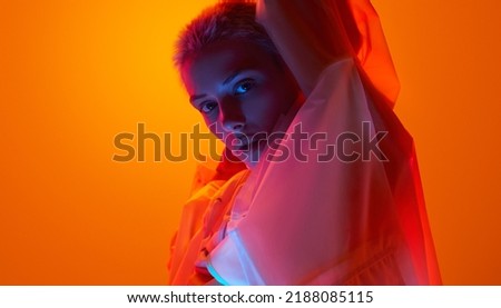 Side view of confident young female model with short blond hair in raincoat, standing against orange background with raised arms and looking at camera in neon light