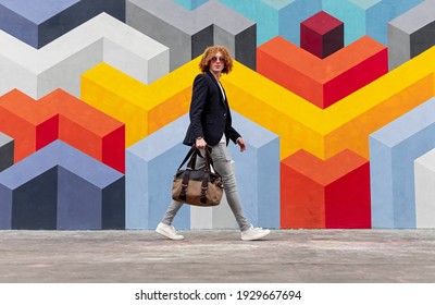 Side view of confident young male with curly red hair in trendy outfit and sunglasses walking on city street, near wall with geometric graffiti and looking at camera - Powered by Shutterstock